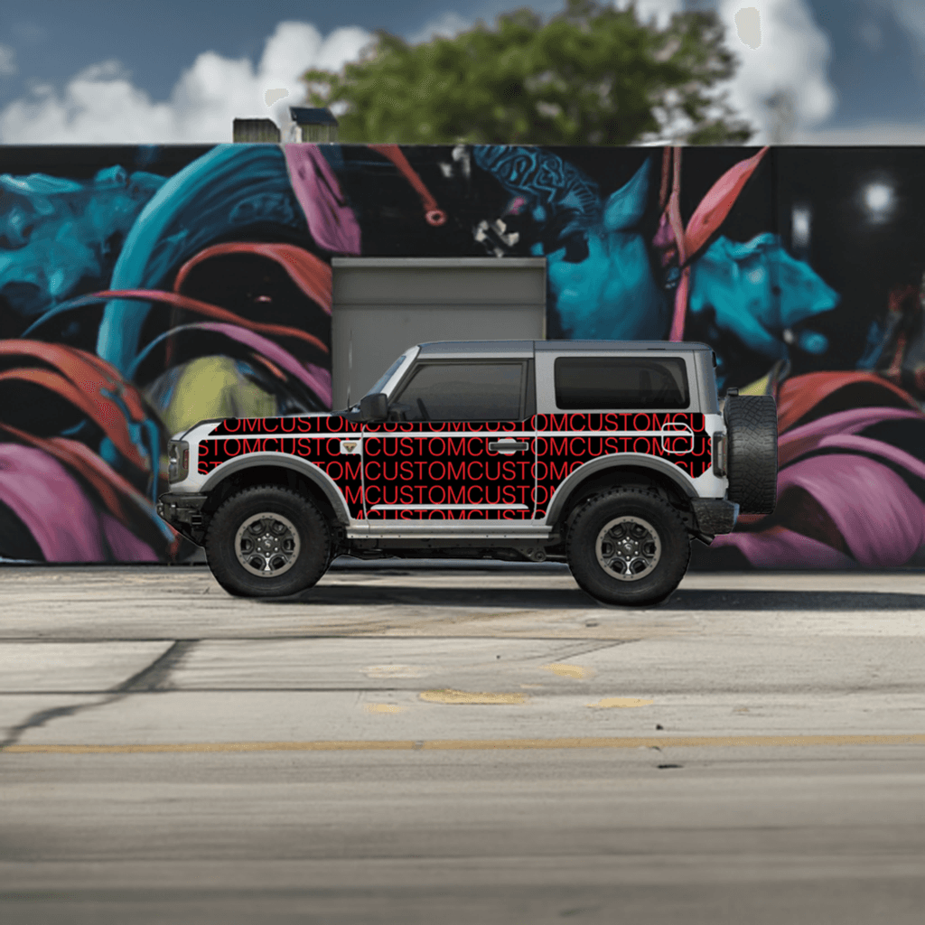 Bronco 2D with custom MEK removable trail armor featuring a bold black and red 'CUSTOM' text design, parked against a vibrant mural background.