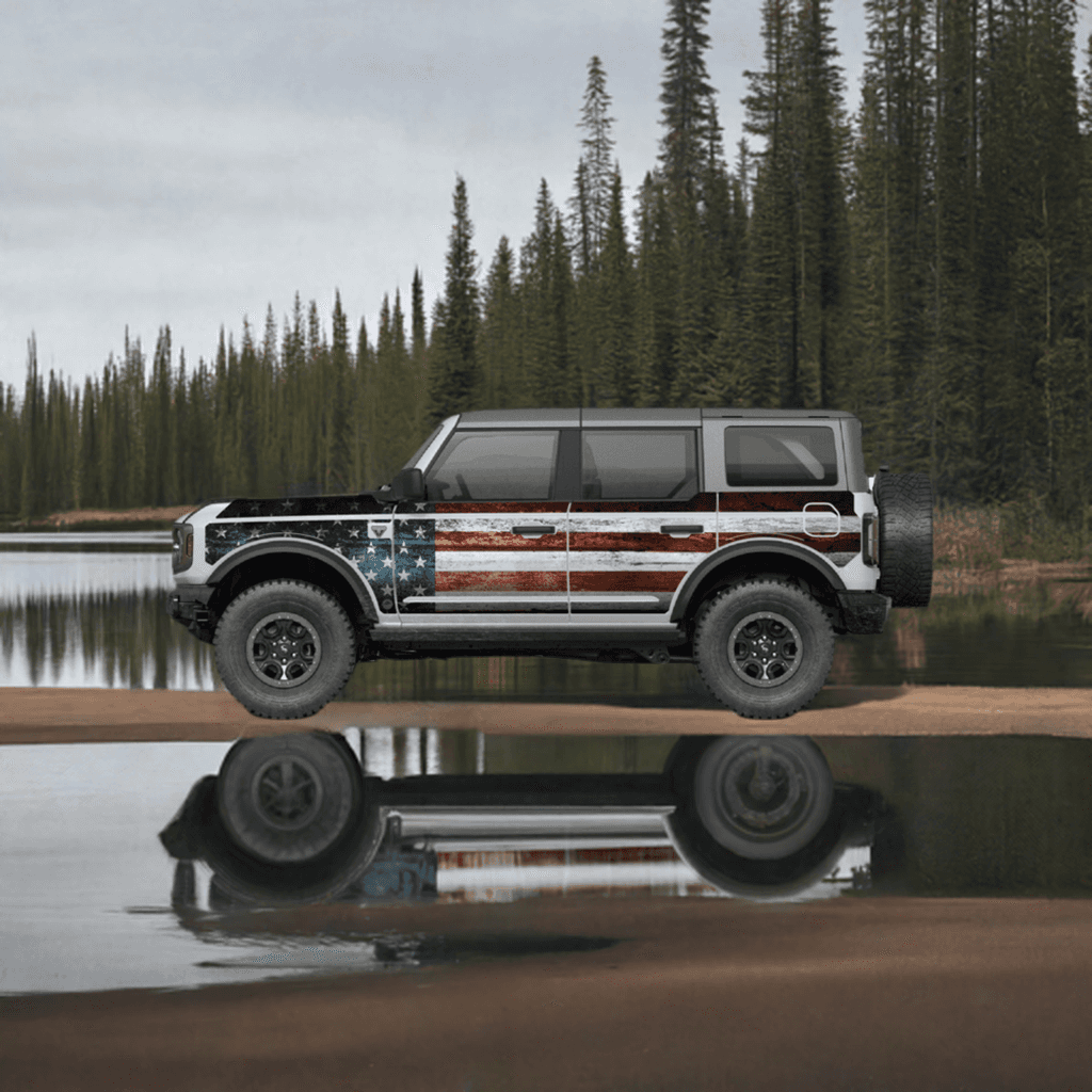 Ford Bronco 4D with MEK removable trail armor featuring an American flag design, parked near a serene lake surrounded by trees.